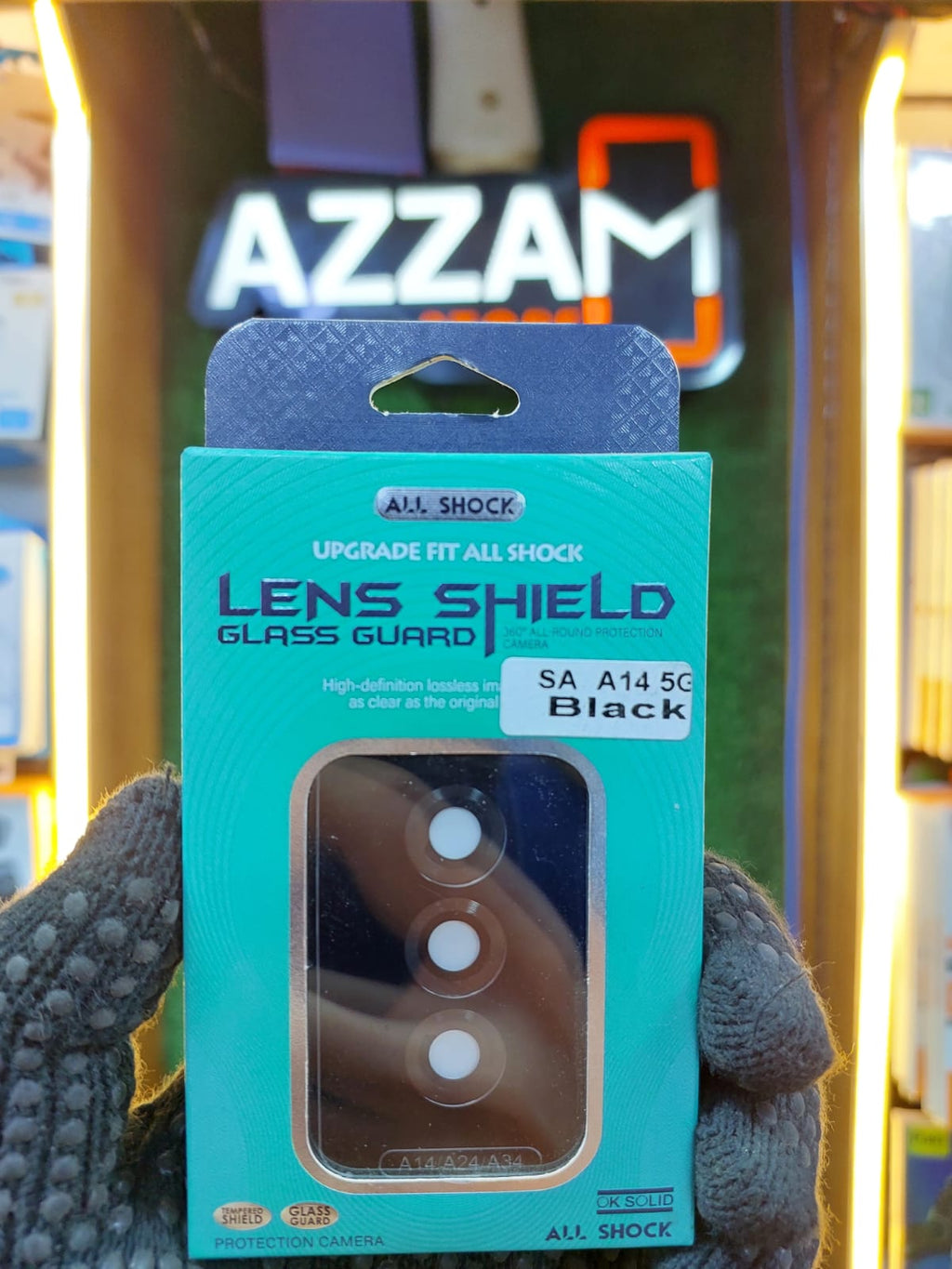 Teal packaging of a lens shield glass guard held by a gloved hand with an Azzam store in the background.